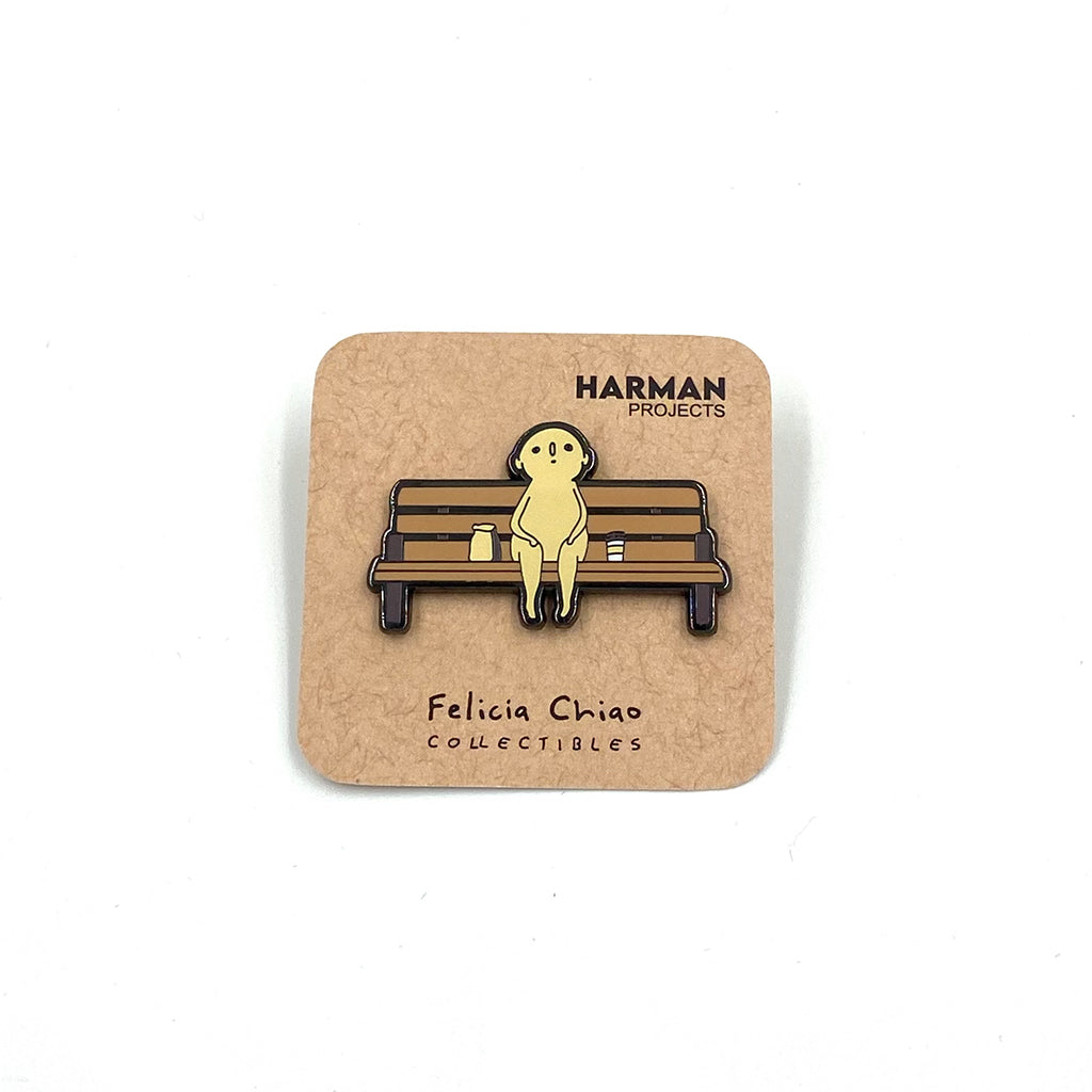 Photo of enamel pin of a figure sitting on a wooden bench with a togo coffee cup to its left and a brown paper bag its right. The pin is on a natural brown backer on a white backdrop. 